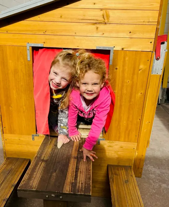 Two Bright Start children smiling through the window of the wooden playhouse